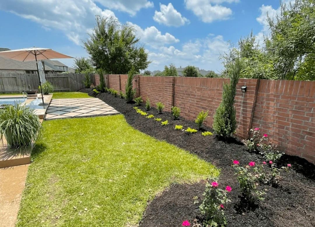 Lush backyard landscape with pool, flower beds, and brick wall under a sunny sky.